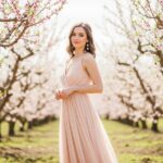 Mladá žena v púdrovo oranžových šatách stojí pod kvitnúcimi broskyňovými stromami.