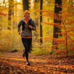 Osoba beží jesenným lesom v vrstvenom oblečení, obklopená spadnutými listami a slnečným svetlom.