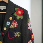 Detailný záber na sako s ručne vyšívanými kvetinovými aplikáciami na drevenom vešiaku.