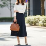 Žena v modrej sukni a bielej blúzke drží koženú kabelku na ulici s prírodným svetlom.