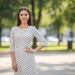 žena v bielych šatách s čiernymi bodkami stojí v parku pri rozmazanej zelenej prírode